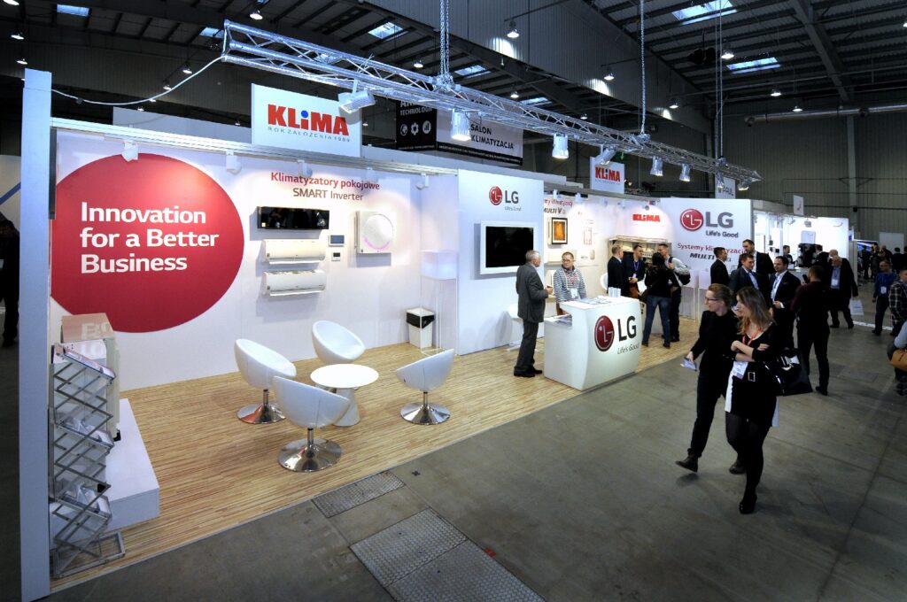Stoisko targowe firmy „Klima na Forum” — białe stoisko drugiej edycji; zawiera autorską kompozycję z modułów Octanorm, czerwone koło z napisem „Innovation for a Better Business”, białe krzesła, ladę LG oraz ludzi nawiązujących kontakt w jasnej hali. projekt i nadzór Tatjana Kobuszewska