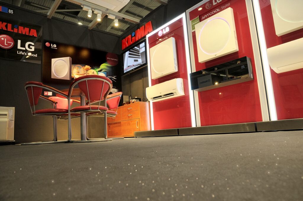 Stoisko wystawowe z logo firm LG i Klima na targach Klima na Forum HVAC — czerwone stoisko autorskie, projekt pracowni TRK System. Urządzenia klimatyzacyjne są wyeksponowane na czerwono-białych panelach, a całość uzupełniają czerwone krzesła z miękkim obiciem oraz szklany stół; całość oświetlają lampy sufitowe.