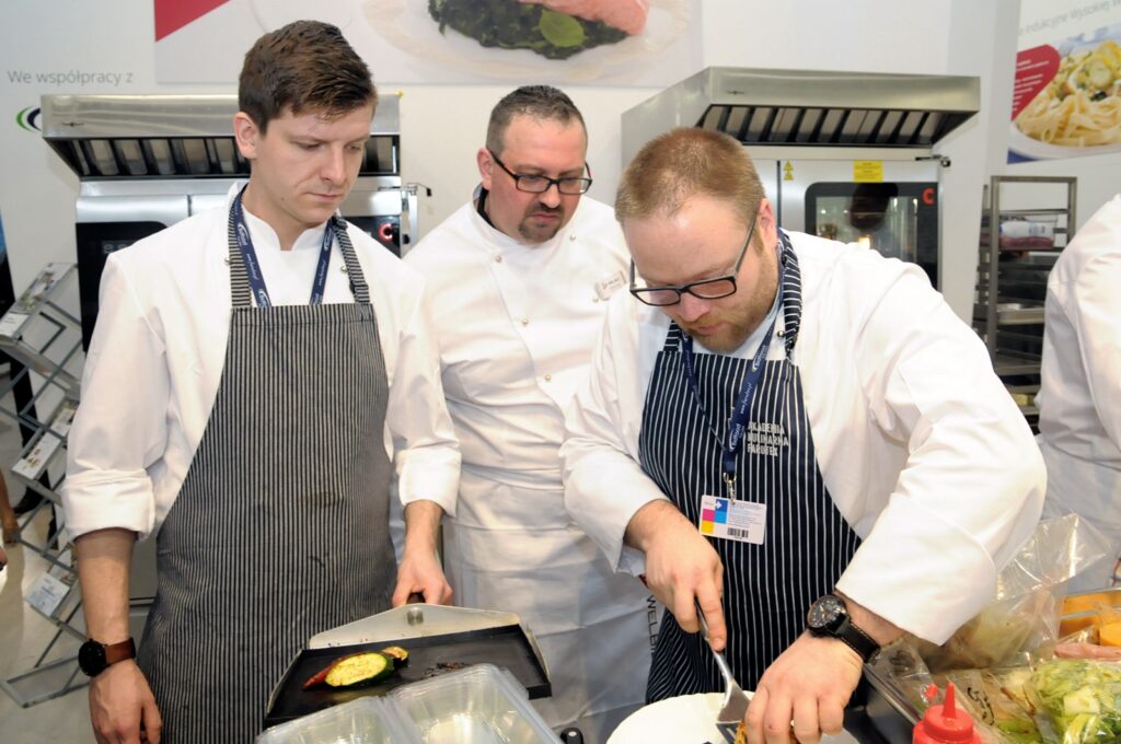 Trzech kucharzy w białych marynarkach, z których dwóch ma na sobie fartuchy w paski, pracuje razem na stoisku Welbilt podczas targów Eurogastro 2017. Jeden z kucharzy kroi potrawę, podczas gdy pozostali obserwują go; w tle widać sprzęt kuchenny i dania na talerzach. projekt i nadzór Tatjana Kobuszewska