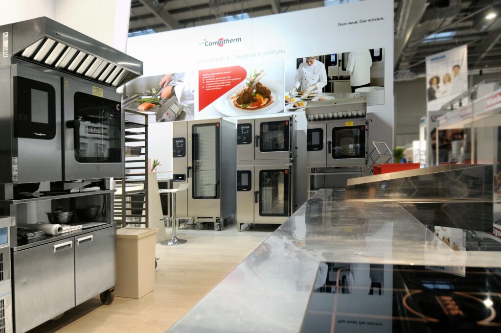 na stoisku Welbilt podczas targów Eurogastro 2017 prezentowane są piekarniki ze stali nierdzewnej, szafy podgrzewające oraz inne urządzenia. są to Convotherm i Merrychef. Na pierwszym planie widoczny jest lśniący blat, a w tle – kucharze oraz duże zdjęcia potraw na banerze wiszącym na ścianie. projekt i nadzór Tatjana Kobuszewska
