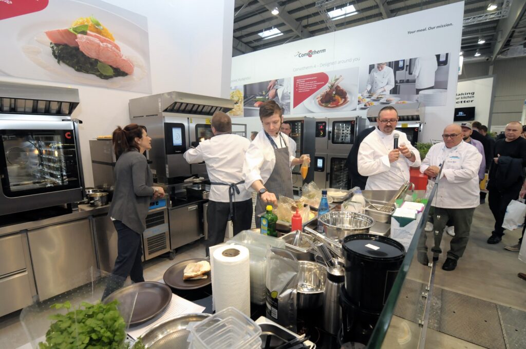W nowoczesnej kuchni na stoisku Welbilt podczas targów Eurogastro 2017 pracuje kilku kucharzy w białych fartuchach, korzystając ze sprzętu ze stali nierdzewnej. Blaty są wypełnione przyborami kuchennymi, miskami i składnikami, a goście obserwują, rozmawiają i oglądają danie wyświetlane na ekranie. projekt i nadzór Tatjana Kobuszewska