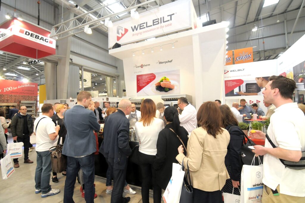 Grupa osób gromadzi się przy stoisku Welbilt na Eurogastro 2017 pod dużym napisem „Welbilt”. Uczestnicy z torbami stoją twarzą do lady lub strefy pokazowej w tym jasnym, nowoczesnym otoczeniu, pełnym banerów i tętniącym życiem. projekt i nadzór Tatjana Kobuszewska
