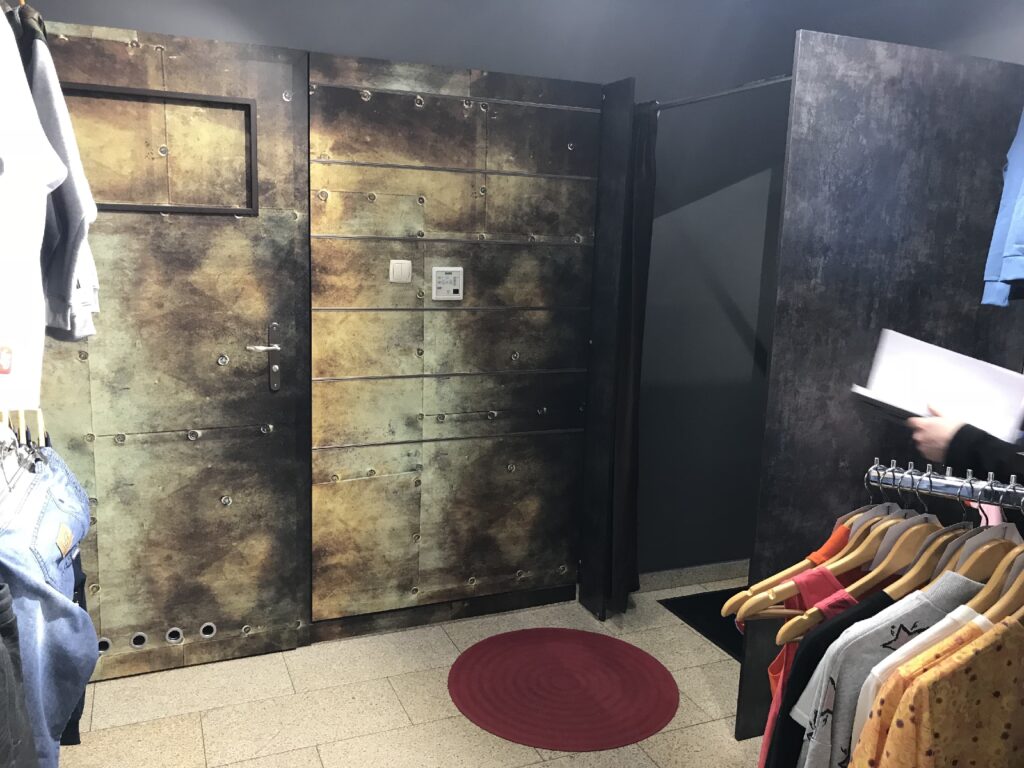 W strefie przymierzalni w sklepie odzieżowym znajduje się ściana w stylu rustykalnym, przypominająca metal, z drzwiami i przełącznikami. Na podłodze wyłożonej płytkami leży czerwony okrągły dywanik. Po prawej i lewej stronie częściowo widoczne są wieszaki z kolorowymi ubraniami oraz osoba trzymająca kartkę papieru. projekt i nadzór Tatjana Kobuszewska