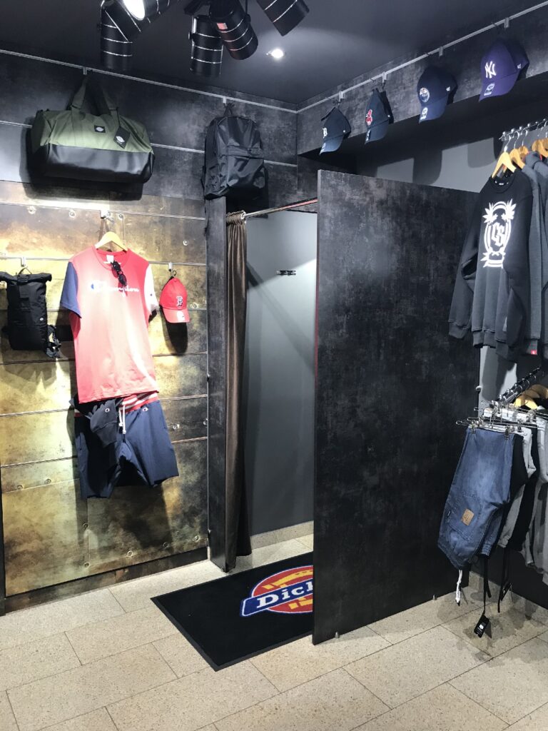 Przymierzalnia z ciemnymi ścianami i industrialnym wystrojem — koncepcja diesel-punkowa dla marki skateboardowej. Czarna zasłona, ubrania w stylu casual, czapki i torby na wystawie. Pod jasnymi światłami, które podkreślają starannie ułożony asortyment, leży mata z napisem „Dickies”. projekt i nadzór Tatjana Kobuszewska
