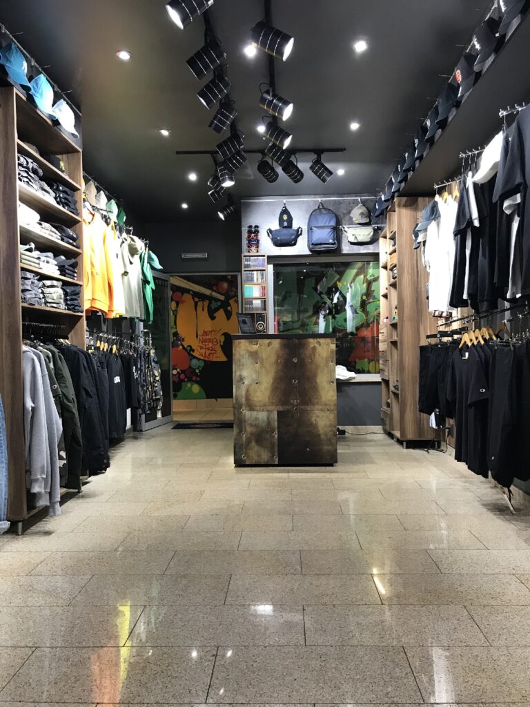 Modny sklep odzieżowy inspirowany sklepem Skate Shop AK-47, z półkami pełnymi czapek, koszulek i kurtek ustawionymi wzdłuż obu ścian. Przestrzeń ta, zaprojektowana przez TRK System w warszawskim podziemnym przejściu, jest nowoczesna i uporządkowana, a za szklanymi drzwiami widnieje kolorowy mural. projekt i nadzór Tatjana Kobuszewska