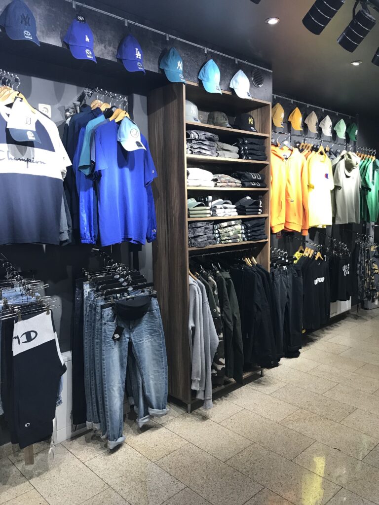 Na wystawie sklepu odzieżowego znajdują się starannie złożone koszule, ułożone w stosy czapki z daszkiem oraz wiszące kurtki, bluzy z kapturem, koszulki i dżinsy w odcieniach niebieskiego, czarnego, pomarańczowego, zielonego i szarego. Oświetlenie punktowe skierowane na produkty podkreśla każdy z nich na podłodze wyłożonej płytkami. projekt i nadzór Tatjana Kobuszewska