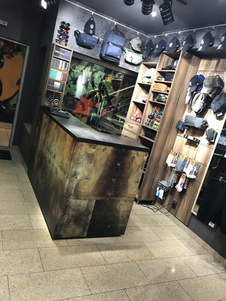 Skate Shop AK-47 — butikowy sklep w warszawskim podziemnym pasażu, z solidną metalową ladą kasową oraz drewnianymi półkami, na których wyeksponowano torby, czapki, koszulki i akcesoria. Reflektory oświetlają czystą, wyłożoną kafelkami podłogę oraz duże lustro na tylnej ścianie. projekt i nadzór Tatjana Kobuszewska