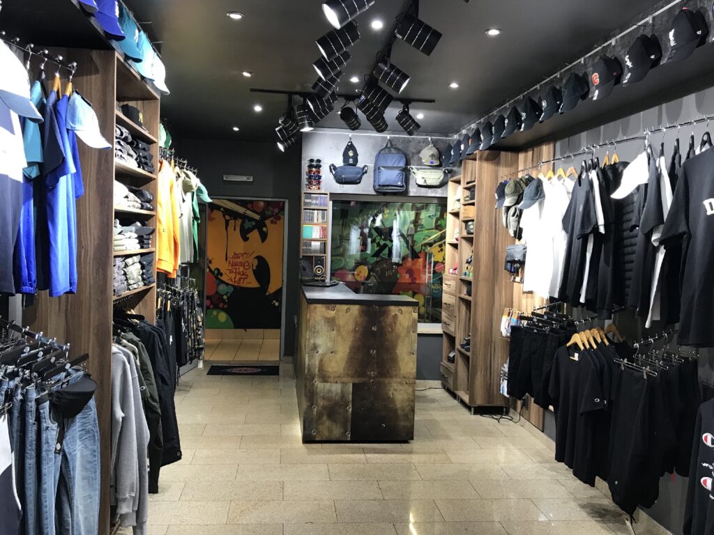 Nowoczesny sklep skateboardowy AK-47 — butik w warszawskim podziemnym pasażu — oferuje starannie ułożone koszulki, kurtki i czapki. Na centralnej ladzie, za którą znajdują się półki, wystawiono plecaki i akcesoria, a jasne oświetlenie podkreśla kolorowe ubrania. projekt i nadzór Tatjana Kobuszewska