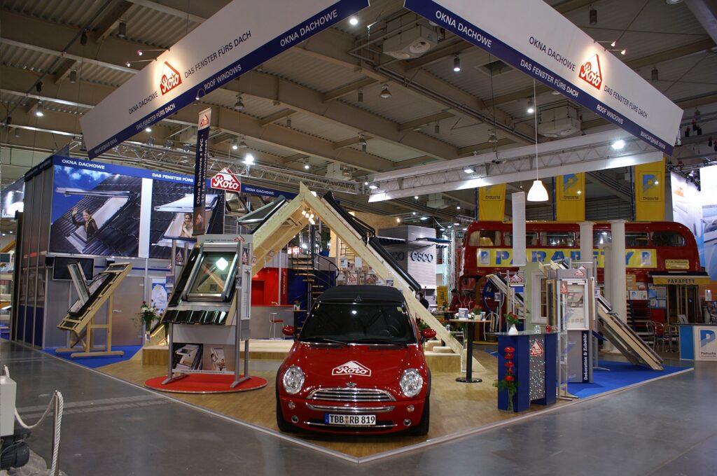 Na stoisku targowym prezentowane są okna dachowe i produkty do świetlików dachowych wraz z zamontowanymi próbkami i plakatami. Na środku stoi czerwony Mini Cooper. Nad głowami gości wyróżnia się duży niebiesko-żółty baner z napisem „Roto” — projektowanie stoisk spaja całość w tej tętniącej życiem, dobrze oświetlonej przestrzeni. projekt i nadzór Tatjana Kobuszewska