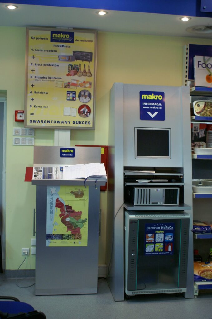 W sklepie stoją obok siebie dwa kioski. Na jednym z nich widnieje duża otwarta książka i mapa. Drugi to cyfrowy kiosk informacyjny z ekranem. Za nimi na tablicy ściennej przedstawiono etapy prowadzące do sukcesu, z tekstem i obrazami inspirowanymi publikacją „Nowoczesna kuchnia komercyjna z rozmieszczonymi Krytycznymi Punktami Kontroli HACCP — projekt technologiczny”. projekt i nadzór Tatjana Kobuszewska