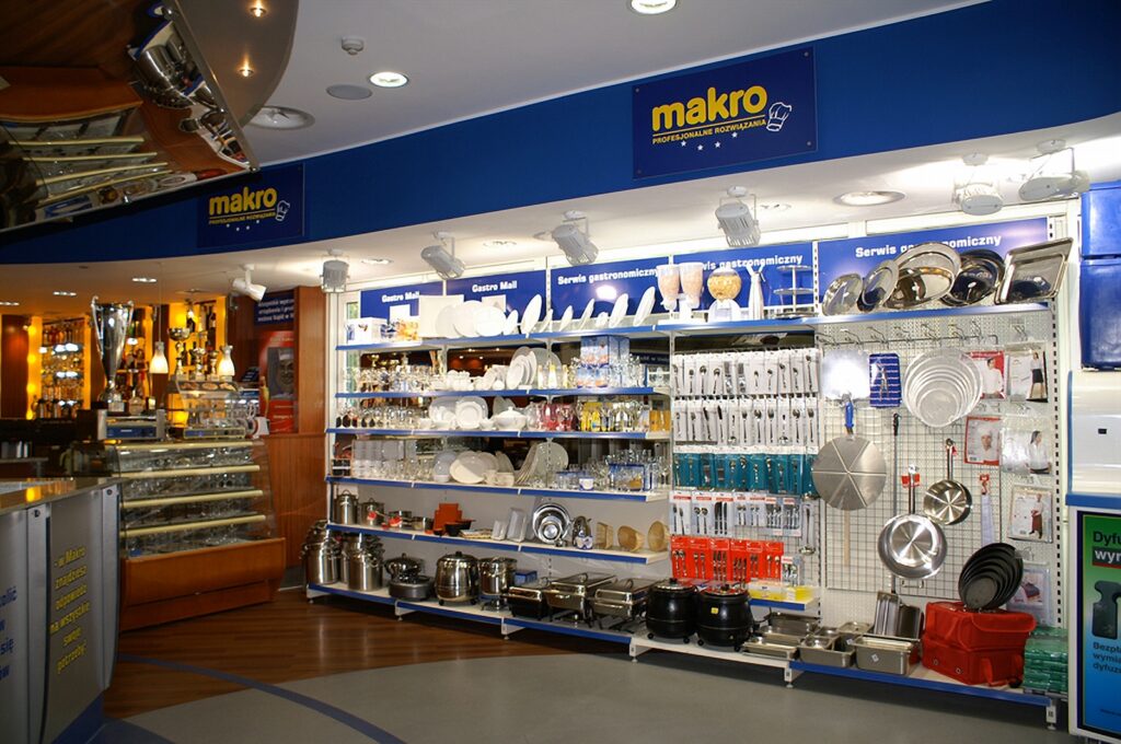 Aleja sklepowa z niebieskimi tablicami z napisem „makro”. Na półkach wystawione są artykuły kuchenne: garnki, patelnie, talerze, szklanki, sztućce oraz akcesoria kuchenne idealne do nowoczesnej kuchni komercyjnej z rozmieszczonymi krytycznymi punktami kontroli HACCP — projekt technologiczny Tatjany Kobuszewskiej. Podłoga jest szara, a ściany mają. projekt i nadzór Tatjana Kobuszewska