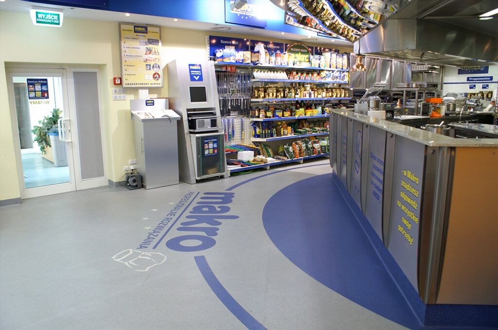 Nowoczesne wnętrze hurtowni z eleganckimi ladami, logo Makro na podłodze, półkami wypełnionymi produktami spożywczymi oraz metalowym wyposażeniem kuchennym po prawej stronie — część nowoczesnej kuchni komercyjnej z rozmieszczonymi krytycznymi punktami kontroli HACCP — projekt technologiczny Tatjany Kobuszewskiej, zrealizowany przy użyciu innowacyjnego systemu TRK. Samodzielny. projekt i nadzór Tatjana Kobuszewska