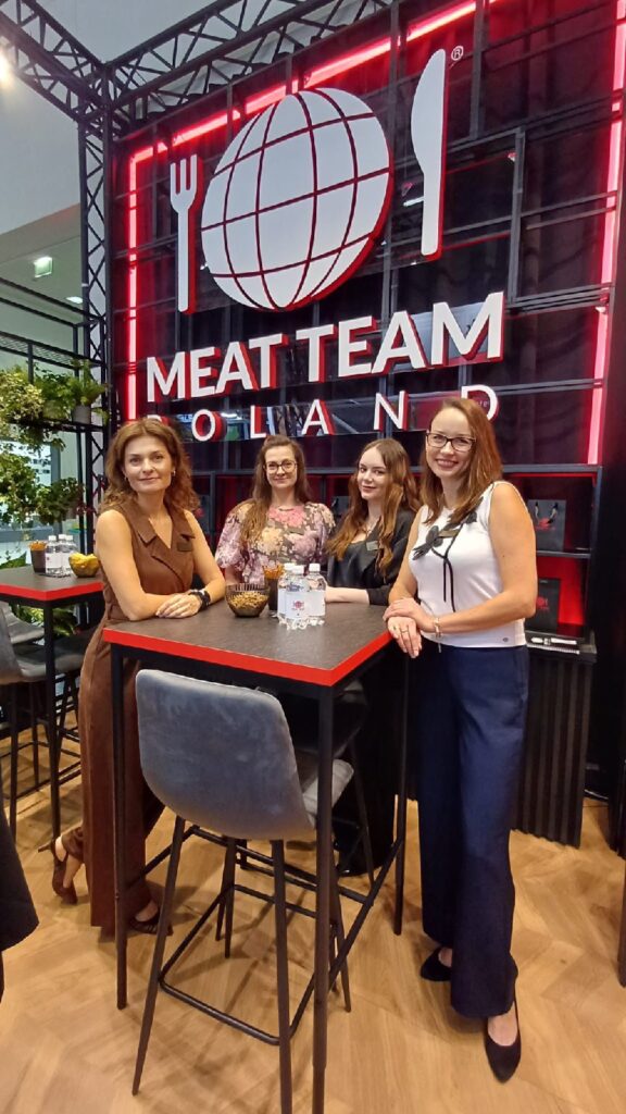 Cztery kobiety stoją z uśmiechem wokół wysokiego stołu podczas imprezy organizowanej w pomieszczeniu. Za nimi widnieje duży napis „MEAT TEAM POLAND” z globusem, widelcem i nożem. Wystrój jest nowoczesny – czarne elementy dekoracyjne, zielone rośliny i drewniana podłoga. projekt i nadzór Tatjana Kobuszewska