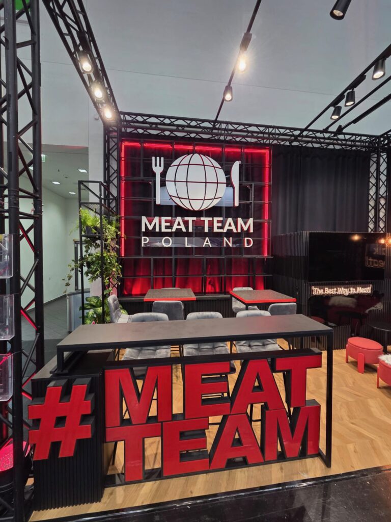 Nowoczesne stoisko wystawowe charakteryzuje się wyrazistym napisem „MEAT TEAM” z przodu, czarnym stołem z szarymi stołkami oraz dużym logo „MEAT TEAM POLAND” umieszczonym powyżej. W przestrzeni dominuje czerwono-czarna kolorystyka, a po lewej stronie widoczne są reflektory punktowe oraz fragment zielonych roślin. projekt i nadzór Tatjana Kobuszewska
