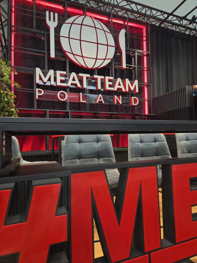 Czerwono-biały napis „MEAT TEAM POLAND” z symbolami globu, widelca i noża na ścianie ekspozycyjnej stoiska targowego Meat Team podczas targów Anuga w Kolonii. Z przodu widoczne są niebiesko-szare wyściełane krzesła, a za nimi czarna lada. Na pierwszym planie widoczne są duże czerwone litery „ME”. W tle widać industrialno-nowoczesny wystrój z metalowymi belkami i roślinami. projekt i nadzór Tatjana Kobuszewska