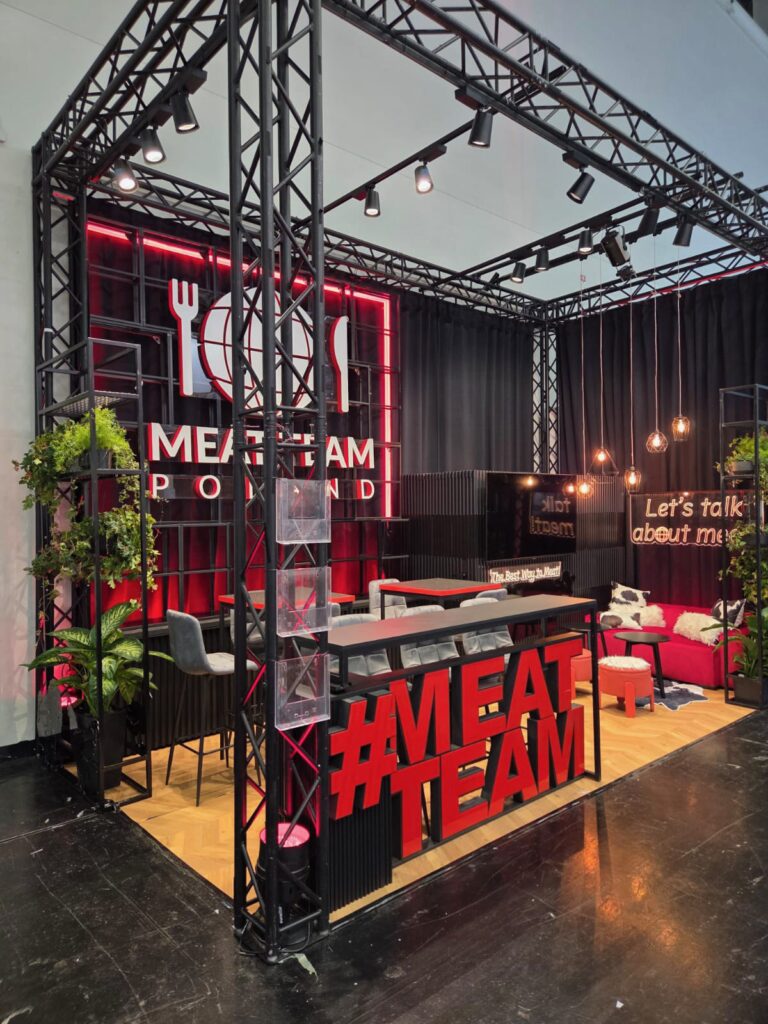 Nowoczesne stoisko wystawowe wyróżnia się dużym napisem „MEAT TEAM POLAND” z motywami zastawy stołowej, czerwono-czarną aranżacją, stołami, krzesłami, czerwoną sofą, roślinami doniczkowymi oraz widocznymi czerwonymi literami „MEAT TEAM” z przodu. Ciepłe lampy wiszące nadają całości przytulną atmosferę. projekt i nadzór Tatjana Kobuszewska