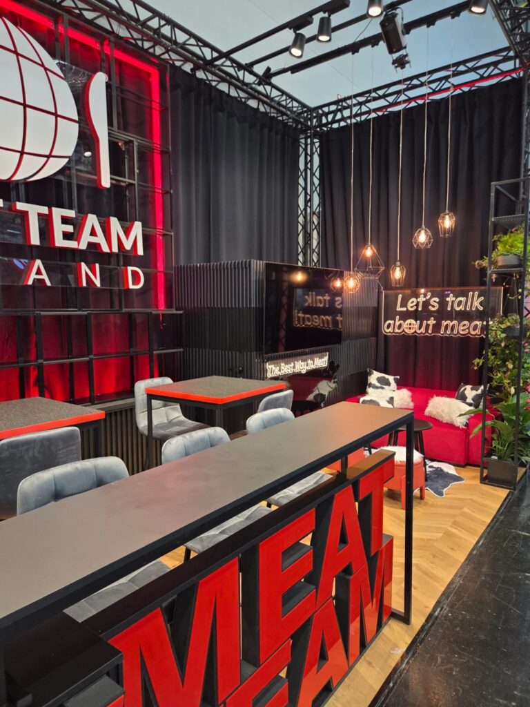 Nowoczesna loża z czerwonym napisem „MEAT TEAM”, wysokimi stołami, szarymi krzesłami, czarnymi zasłonami, wiszącymi lampami, neonowym napisem „Lets talk about meat”, dywanikiem ze skóry bydlęcej, sztucznymi roślinami oraz przytulną strefą wypoczynkową, tworząca przyjazną, nowoczesną atmosferę. projekt i nadzór Tatjana Kobuszewska
