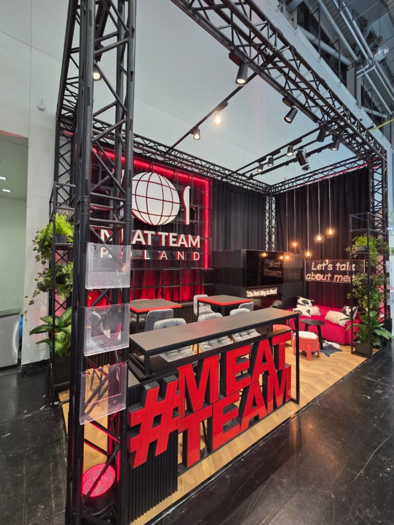 Nowoczesne stoisko wystawowe z dużym czerwonym napisem „MEATTEAM” z przodu, czarną ramą, roślinnością, miejscami do wypoczynku oraz neonowymi napisami „MEAT TEAM POLAND” i „Lets talk about meat”. Aranżacja jest stylowa, z drewnianą podłogą i przytulnym oświetleniem. projekt i nadzór Tatjana Kobuszewska