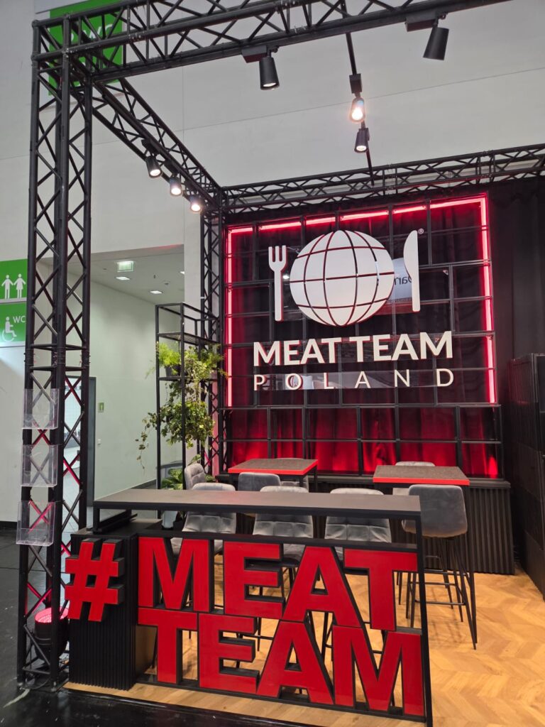 Stoisko wystawowe z czarną ramą, neonowym napisem w kolorze czerwonym „MEAT TEAM POLAND” oraz dużym napisem „MEAT TEAM” z przodu. Po lewej stronie znajdują się szare krzesła, stół, lampy sufitowe oraz roślina. W tle widoczna jest biała ściana i zielony znak wskazujący toaletę. projekt i nadzór Tatjana Kobuszewska