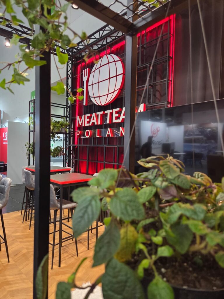 Na czerwono-białym szyldzie widnieje napis „MEAT TEAM POLAND” wraz z logo przedstawiającym globus i widelec. Szyld jest otoczony czarnymi metalowymi konstrukcjami. Na pierwszym planie widok częściowo zasłaniają bujne, zielone rośliny. W tle widać nowoczesne krzesła i stoły. projekt i nadzór Tatjana Kobuszewska