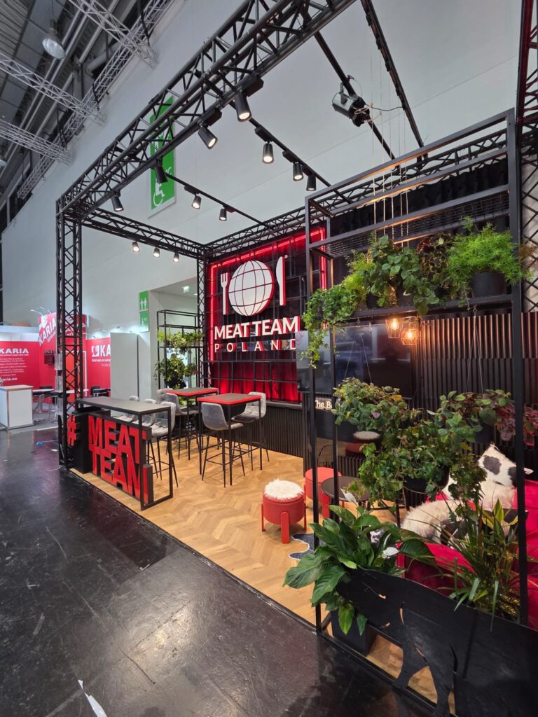 Stoisko targowe firmy MEAT TEAM POLAND z czarną metalową konstrukcją, podświetlonym na czerwono napisem, stołem jadalnym, krzesłami oraz zielonymi roślinami zdobiącymi przestrzeń. Podłoga ma wzór imitujący drewno, a w całej tej stylowej aranżacji widoczne są czerwone i białe akcenty. projekt i nadzór Tatjana Kobuszewska
