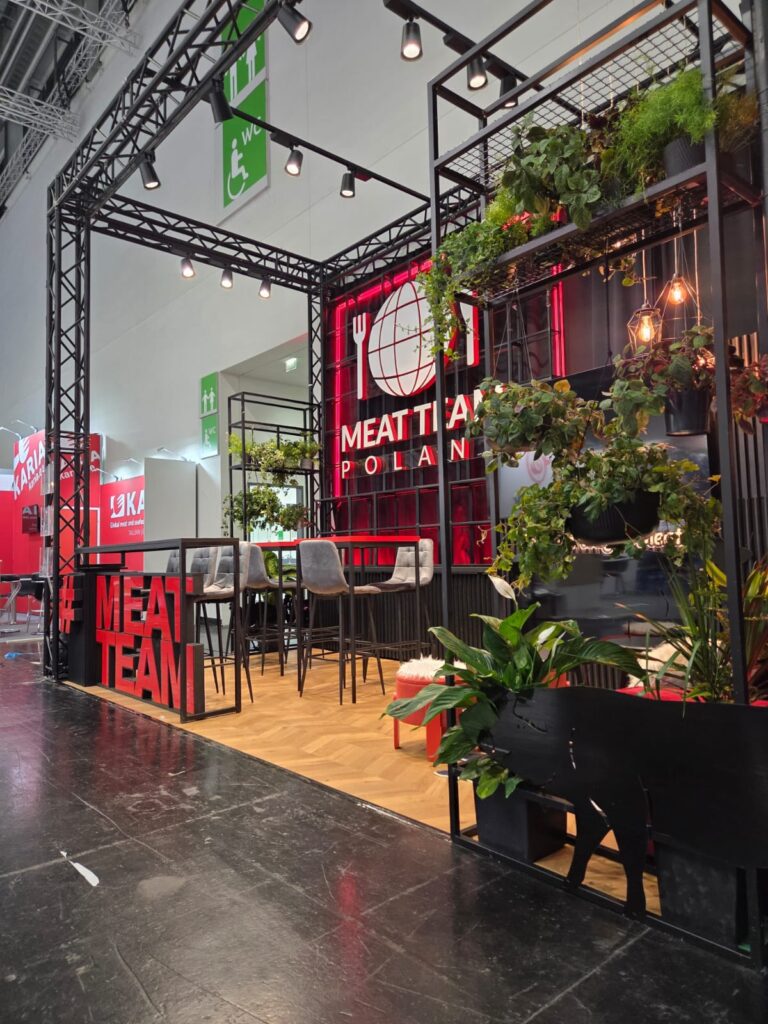 Nowoczesne stoisko targowe z napisem „Meat Team Poland”, czarną metalową konstrukcją, wiszącymi roślinami i drewnianą podłogą. Widoczna jest czerwono-biała identyfikacja wizualna, a wewnątrz znajdują się krzesła i stoły. Z przodu stoiska stoi duży czerwony napis „MEATTEAM”. Nad stoiskiem wiszą znaki wskazujące wyjście. projekt i nadzór Tatjana Kobuszewska