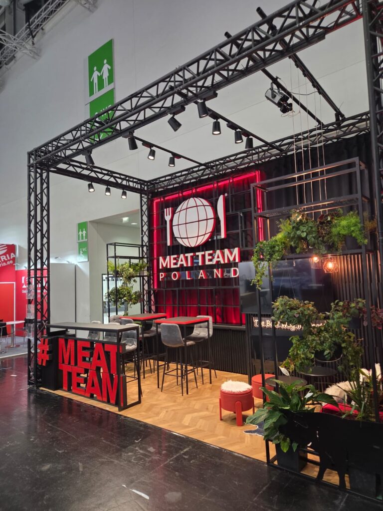 Stoisko targowe z napisem „MEAT TEAM POLAND”, czerwono-białym logo w kształcie globu, czarną metalową ramą, roślinami, wiszącymi lampami, wysokimi stołkami i stołami. Przestrzeń zdobi czerwony napis „MEAT TEAM” oraz rośliny. Na białej ścianie powyżej widoczne są znaki wyjścia awaryjnego. projekt i nadzór Tatjana Kobuszewska