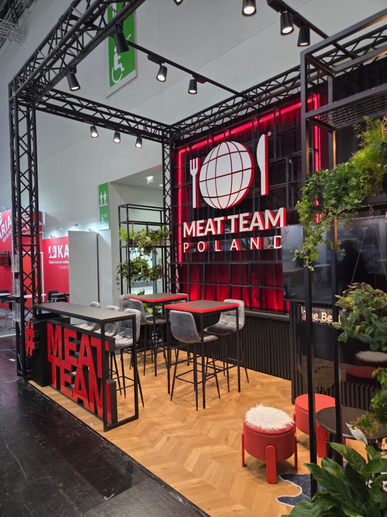 Nowoczesne stoisko targowe firmy MEAT TEAM POLAND („stoisko targowe Meat Team na targach Anuga w Kolonii”) charakteryzuje się efektowną czerwono-czarną kolorystyką, logo w kształcie globusa z motywem sztućców, wysokimi stołami ze stołkami, roślinami oraz reflektorami punktowymi zamontowanymi nad głowami. Hashtagi i rośliny stanowią elementy dekoracyjne, a parkiet dopełnia ten nowoczesny wygląd. projekt i nadzór Tatjana Kobuszewska