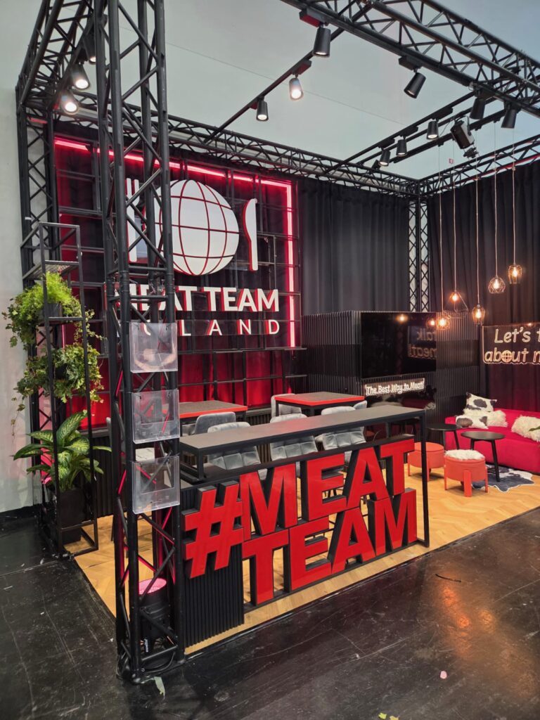 Nowoczesne stoisko targowe „MEAT TEAM” wyróżnia się dużym napisem w kolorze czerwonym, ladą, stołkami, roślinami, kanapami oraz logo przedstawiającym globus, widelec i nóż na tylnej ścianie. W stylowej aranżacji dominują kolory czerwony i czarny — stoisko przyciąga wzrok na targach Anuga w Koeln Messe. projekt i nadzór Tatjana Kobuszewska