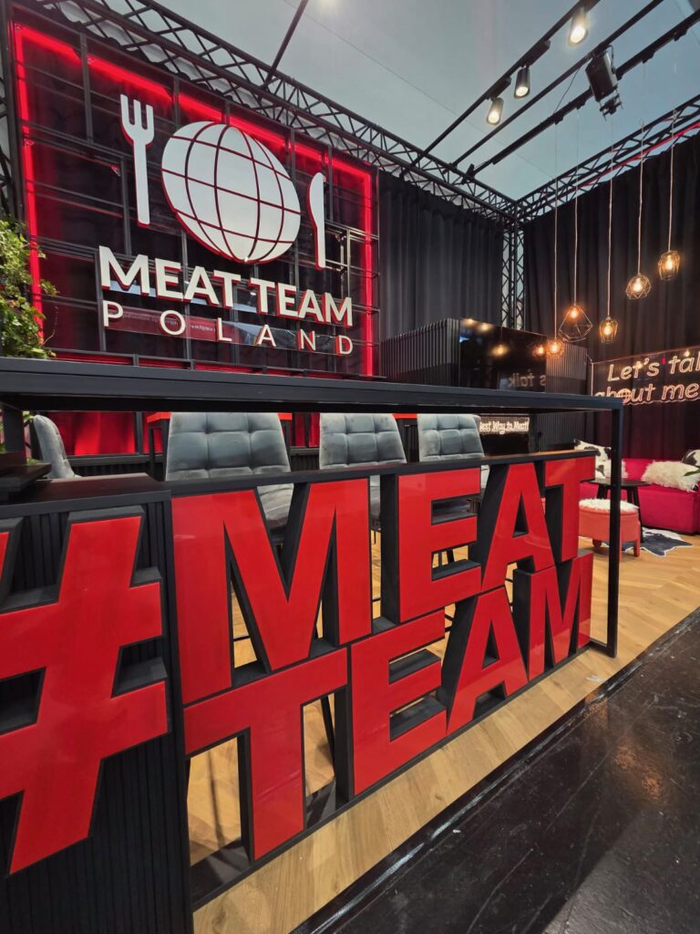 Nowoczesne stoisko z dużym czerwonym napisem „MEATTEAM” z przodu. Z tyłu szare krzesła i czarny stół. Na tylnej ścianie podświetlone na czerwono logo „MEAT TEAM POLAND” z globusem i przyborami kuchennymi. Po prawej stronie dekoracyjne oświetlenie i strefa wypoczynkowa. Miejska, stylowa aranżacja targowa. projekt i nadzór Tatjana Kobuszewska