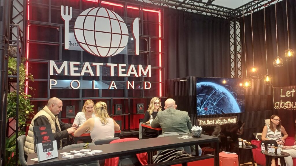 Sześć osób siedzi przy stolikach w loży, na której ścianie widnieje napis „Meat Team Poland” wykonany jasnymi neonowymi literami. Loża urządzona jest w czarno-czerwonej kolorystyce; znajduje się w niej duże logo w kształcie globusa, ekran telewizyjny z obrazami potraw, półki z produktami oraz materiały promocyjne na stolikach. projekt i nadzór Tatjana Kobuszewska