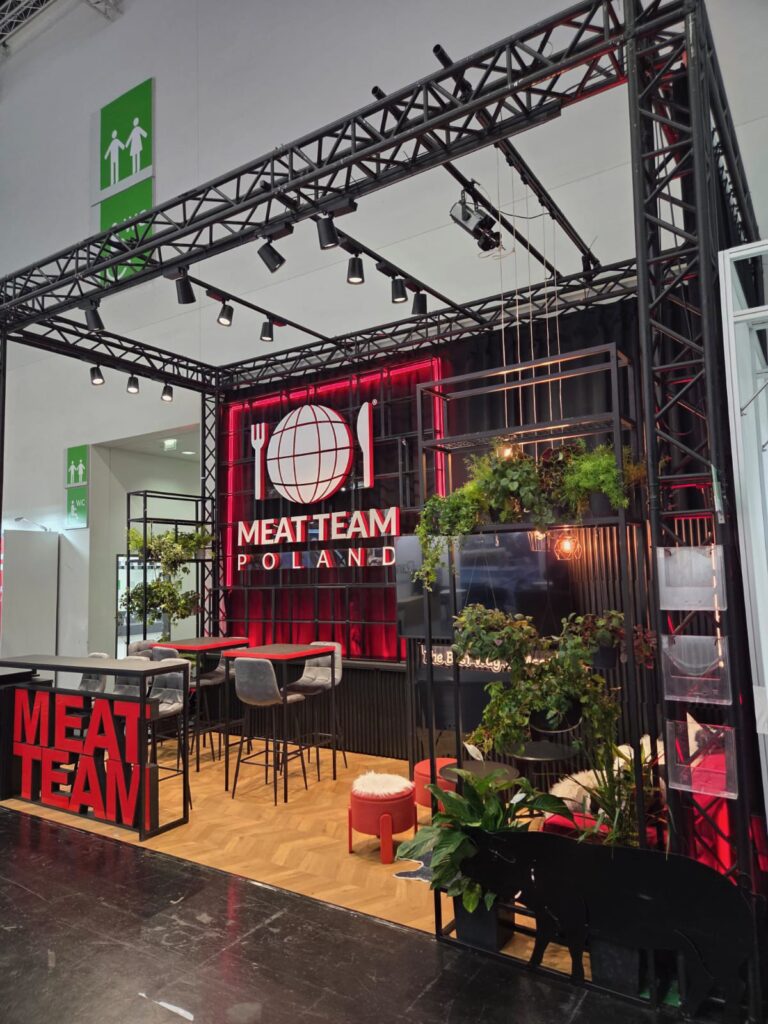 Stoisko targowe w stylu industrialnym z logo MEAT TEAM POLAND, czarną metalową ramą, czerwonym oświetleniem, stołami barowymi i stołkami, ławką z imitacji trawy, roślinami doniczkowymi oraz dekoracją w kształcie świnki. Na białej ścianie za stoiskiem wiszą zielone znaki wyjścia awaryjnego. projekt i nadzór Tatjana Kobuszewska