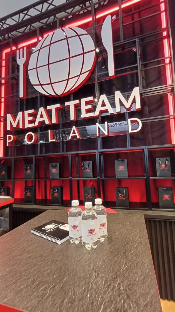Duży napis „MEAT TEAM POLAND” z logo przedstawiającym globus, widelec i nóż. Na blacie leżą trzy butelki wody i czarny notatnik z tym logo. W tle widoczne są czarne regały i czerwone oświetlenie. Nowoczesny, odważny projekt. projekt i nadzór Tatjana Kobuszewska