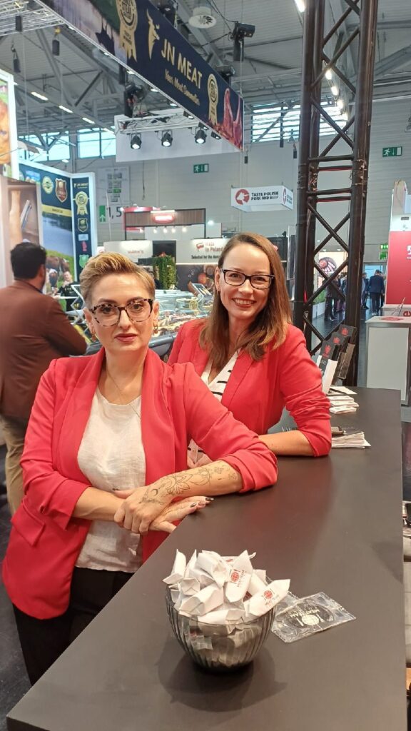 Dwie kobiety w identycznych czerwonych marynarkach i okularach stoją przy ladzie i uśmiechają się. Jedna z nich pochyla się do przodu ze skrzyżowanymi rękami, a druga stoi za nią. Na ladzie stoi miska z złożonymi kartkami. W tle widać tętniące życiem targi w pomieszczeniu z różnymi stoiskami. projekt i nadzór Tatjana Kobuszewska