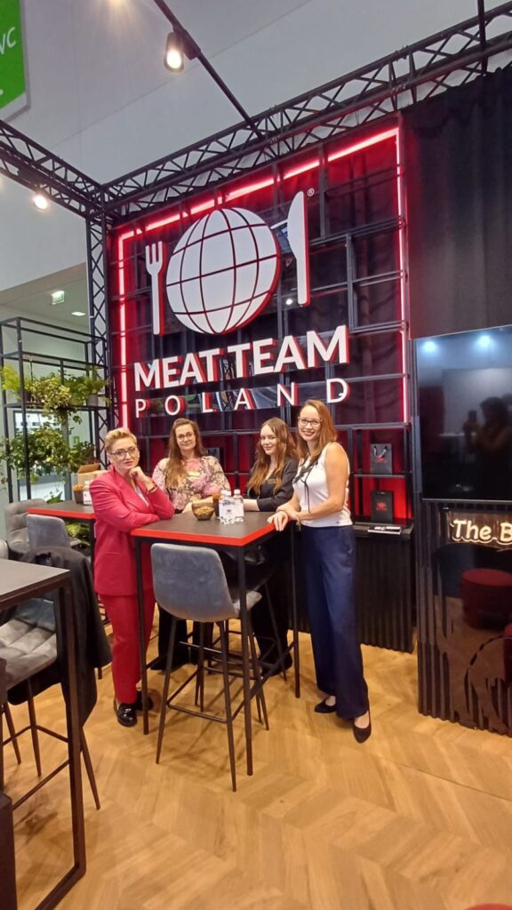 Cztery kobiety stoją i uśmiechają się przy wysokim stole w krytym stoisku targowym, nad którym widnieje duży neon z napisem „Meat Team Poland” oraz logo przedstawiające globus, widelec i nóż. Stoisko Meat Team wyróżnia się efektowną czarno-czerwoną aranżacją z drewnianą podłogą, przyciągając uwagę na targach Anuga w Koeln Messe. projekt i nadzór Tatjana Kobuszewska