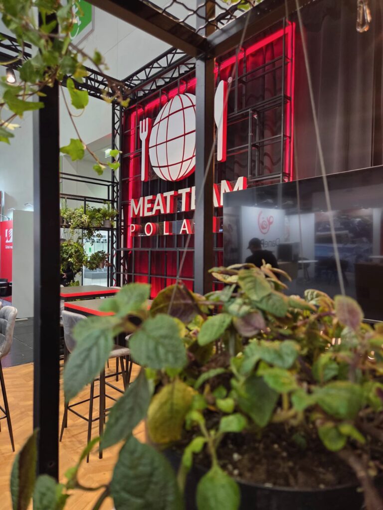 Zbliżenie na wiszące na pierwszym planie zielone rośliny liściaste, a w tle widoczny czerwono-biały napis „Meat Team Poland” oraz nowoczesne czarne metalowe meble na wystawie lub targach organizowanych w pomieszczeniu. projekt i nadzór Tatjana Kobuszewska