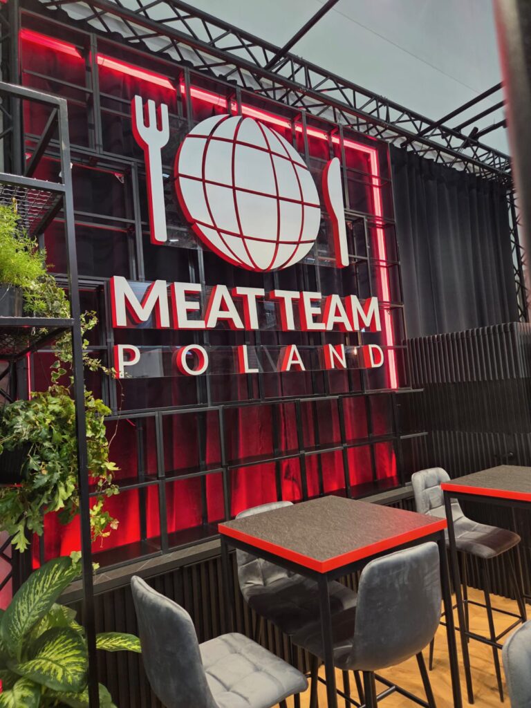 Nowoczesne wnętrze restauracji z świecącym czerwono-białym napisem „MEAT TEAM POLAND” umieszczonym pod logo przedstawiającym globus, widelec i nóż. W środku znajdują się czarne stoły z szarymi krzesłami, a przestrzeń zdobią zielone rośliny. Tło tworzą czarne ściany i zasłony. projekt i nadzór Tatjana Kobuszewska