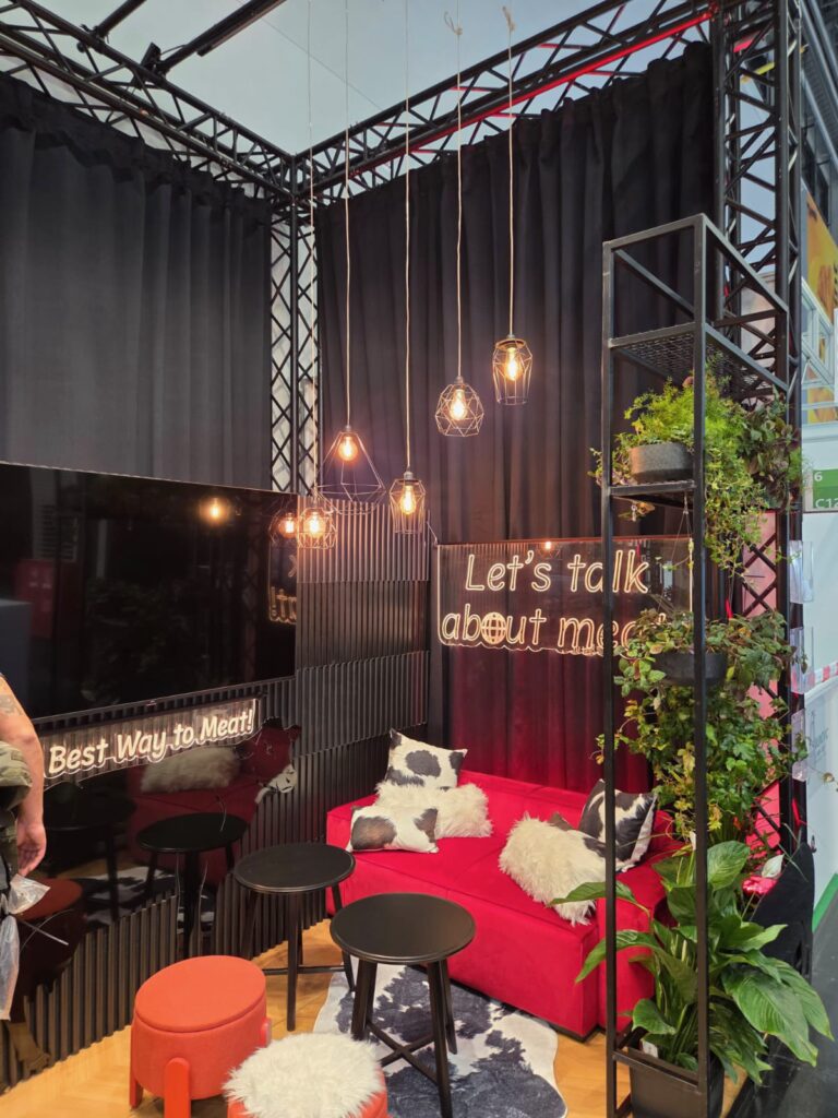 Przytulny salon z czerwoną sofą, czarnymi zasłonami, wiszącymi lampami, pluszowymi poduszkami, małymi stolikami i zielonymi roślinami. Na neonowych napisach widnieją hasła: „Best Way to Meat” i „Lets talk about meat”. Przestrzeń sprawia wrażenie nowoczesnej i zachęcającej. projekt i nadzór Tatjana Kobuszewska