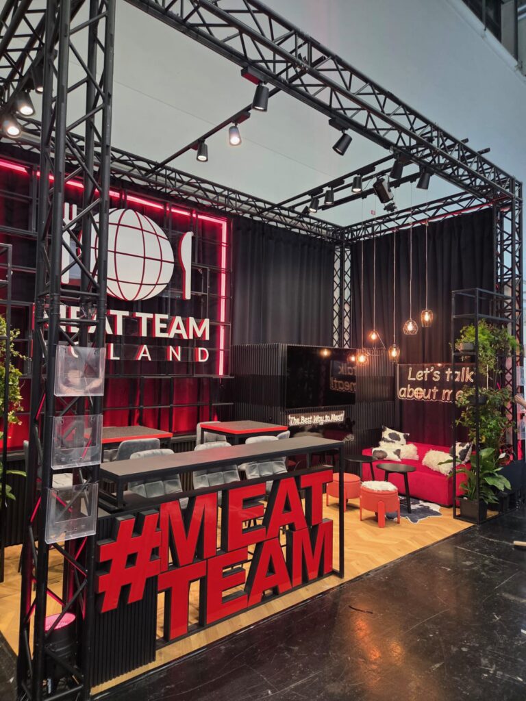 Nowoczesne stoisko targowe z czerwonym neonowym napisem „MEAT TEAM”, ekspozycją hashtagów, czarnymi ramami, reflektorami punktowymi, wysokimi stołami i stołkami, pluszowymi czerwonymi sofami, wiszącymi żarówkami, roślinami oraz banerami z napisem „Porozmawiajmy o mięsie”. Elegancka, zachęcająca aranżacja na drewnianej podłodze. projekt i nadzór Tatjana Kobuszewska