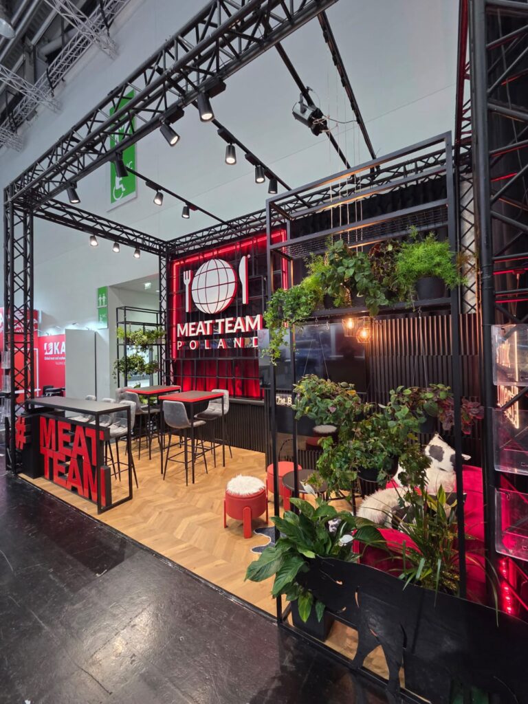 Nowoczesne stoisko targowe zawiera napis „MEAT TEAM POLAND”, czarne metalowe ramy, czerwone akcenty, bujne zielone rośliny, stołki barowe, małe stoliki oraz wiszące lampy. Tło jest w kolorach czarnym i czerwonym, z motywem globusa, logo przedstawiającym przybory kuchenne oraz drewnianą podłogą. projekt i nadzór Tatjana Kobuszewska