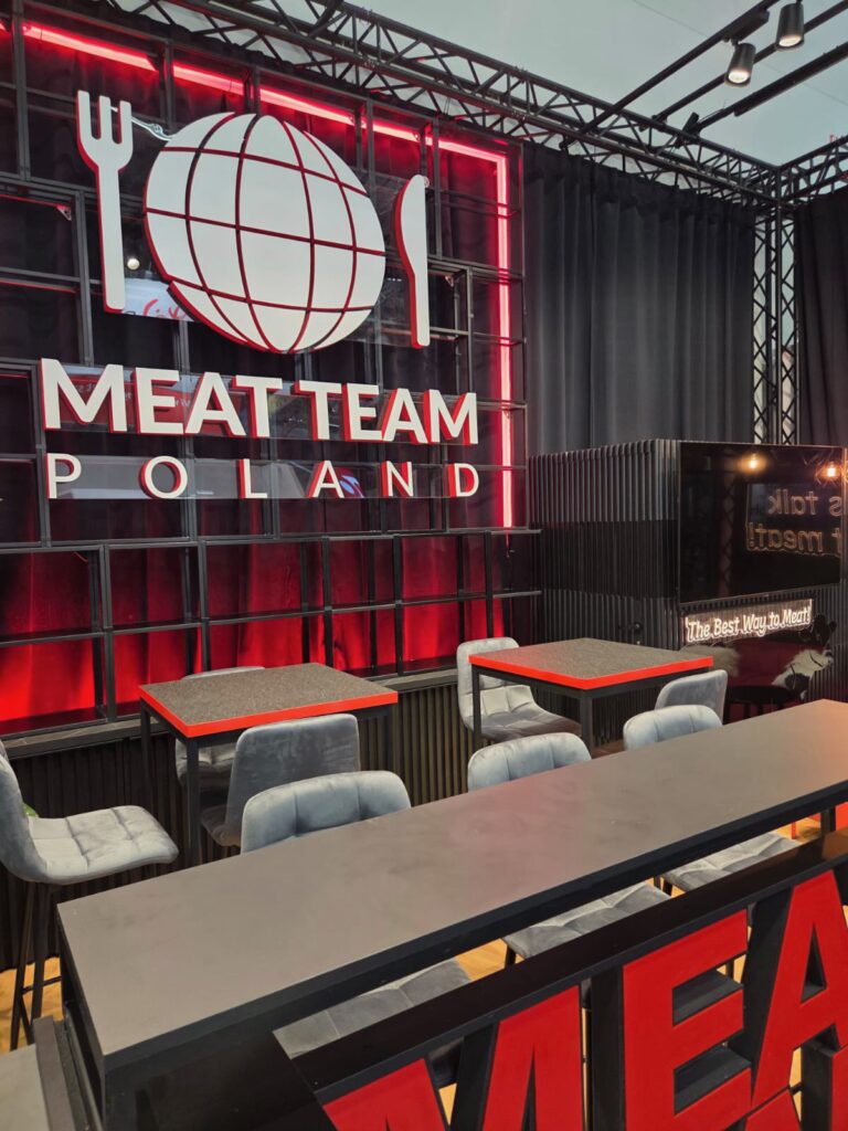 Nowoczesne stoisko firmy MEAT TEAM POLAND z czerwono-białym napisem w stylu neonowym przedstawiającym globus, widelec i nóż. Przed stoiskiem ustawiono szare stoły i krzesła. Na bocznym ekranie widoczny jest slogan „The Best Way to Meat”. projekt i nadzór Tatjana Kobuszewska