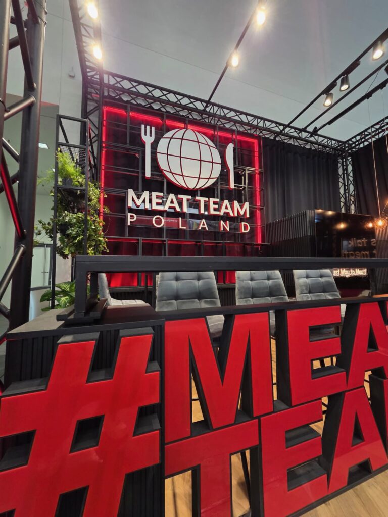 Nowoczesne stoisko wystawowe prezentuje napis „MEAT TEAM POLAND” wykonany pogrubioną czcionką pod globusem z widelcem i nożem. Z przodu stoją czarne krzesła i duży czerwony napis „MEAT”; stoisko zdobią czarne ramki i rośliny. projekt i nadzór Tatjana Kobuszewska