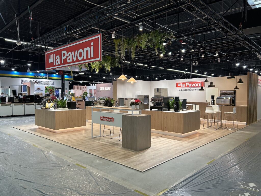 Nowoczesne stoisko targowe firmy La Pavoni składa się z drewnianych lad, zielonych roślin, ekspresów do kawy, lamp wiszących oraz czerwonych napisów „La Pavoni”. Stoisko stoi na podłodze imitującej drewno, otoczonej podłogą pokrytą folią w dużej hali wystawowej. projekt i nadzór Tatjana Kobuszewska