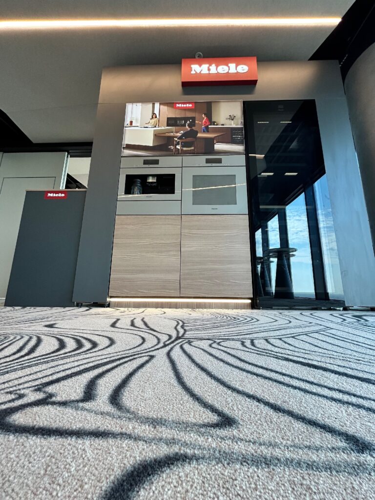 Ujęcie pod niskim kątem prezentującej sprzęt AGD marki Miele z czerwonym napisem „Miele”, wbudowanymi piekarnikami i eleganckimi szafkami. Na podłodze leży jasna wykładzina z ciemnymi, artystycznymi wzorami w kształcie zawijasów. Duże okna po prawej stronie odbijają błękitne niebo i chmury. projekt i nadzór Tatjana Kobuszewska
