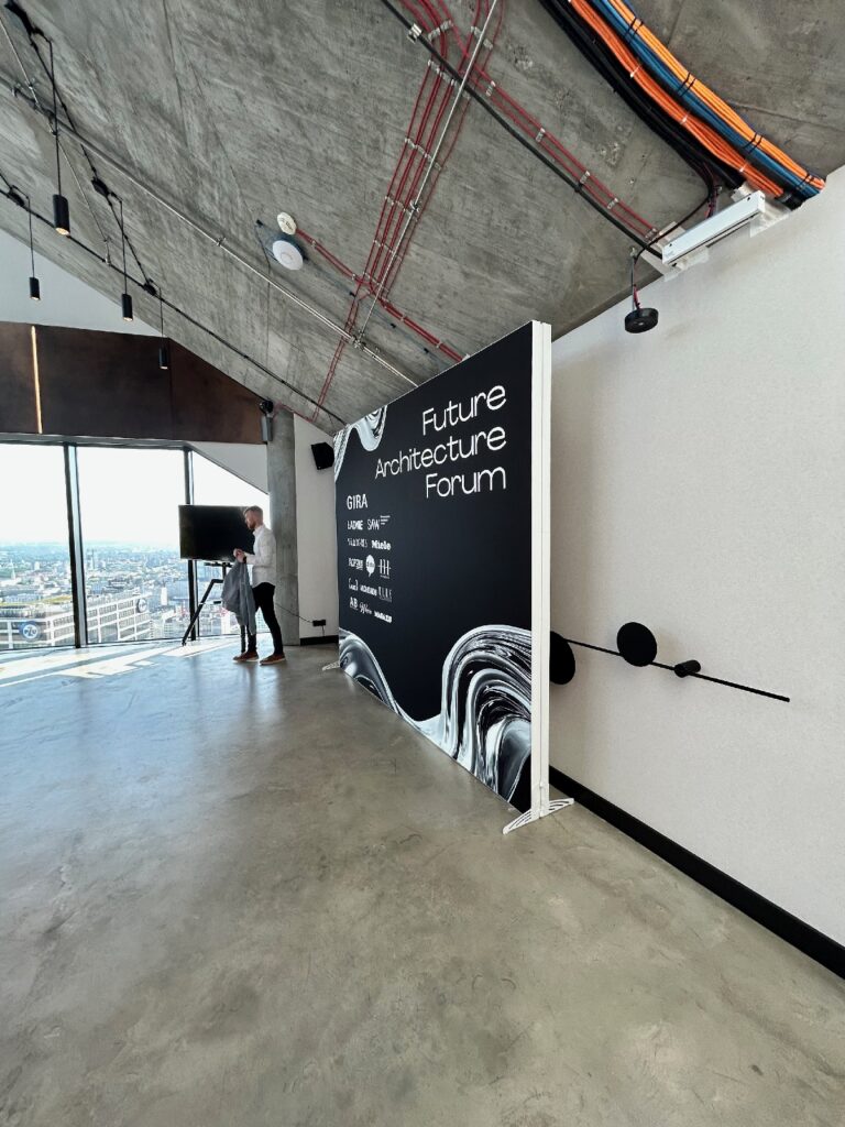 W nowoczesnym pomieszczeniu urządzonym w stylu industrialnym wisi wysoki baner z napisem „Future Architecture Forum”. Obok okien sięgających od podłogi do sufitu, z których rozciąga się widok na miasto, stoi osoba. Wzdłuż betonowego stropu biegną odsłonięte rury i przewody. Widoczny jest telewizor na stoliku. projekt i nadzór Tatjana Kobuszewska