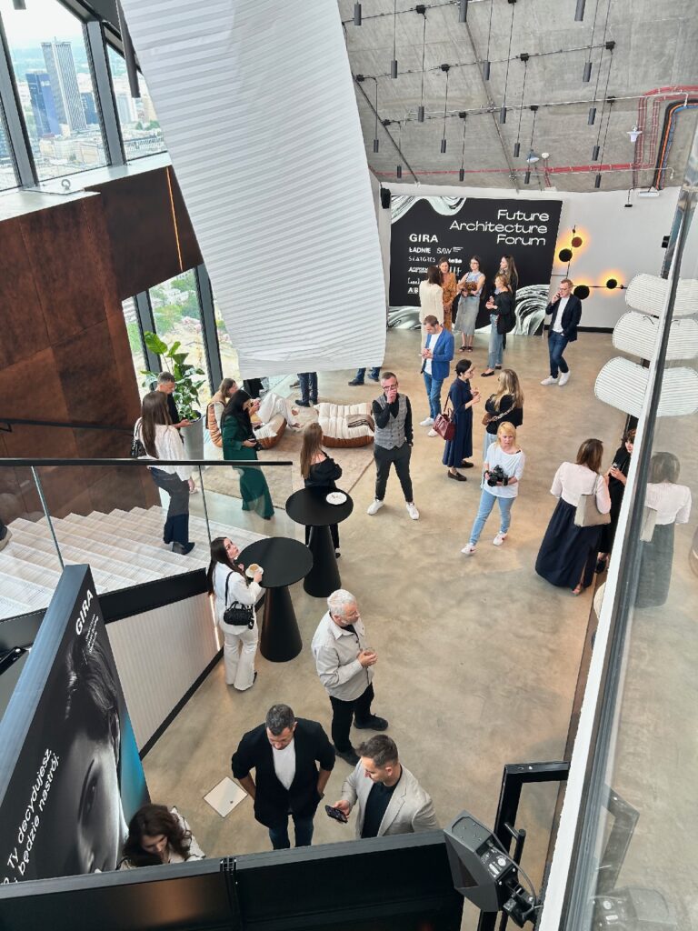 Ludzie przebywa w nowoczesnej przestrzeni eventowej w stylu industrialnym, z betonowymi ścianami i dużymi oknami. Widoczny jest napis „Future Architecture Forum”. Uczestnicy rozmawiają, stoją przy okrągłych stołach i spacerują po dwupoziomowej przestrzeni z białymi schodami. projekt i nadzór Tatjana Kobuszewska