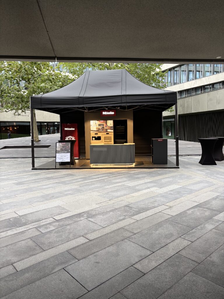 Na wybrukowanym placu w Royal Wilanów stoi czarny namiotowy kiosk z logo firmy Miele. W środku elegancka zabudowa obejmuje jasne drewniane półki, ladę oraz nowoczesne urządzenia. Na zewnątrz znajduje się stojak ekspozycyjny i mały kosz na śmieci, a w tle widać drzewa i współczesne budynki. projekt i nadzór Tatjana Kobuszewska