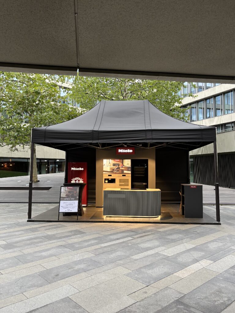 W czarnym, zadaszonym stoisku typu pop-up z napisem „Miele” prezentowane są urządzenia kuchenne w ramach eleganckiej zabudowy. Znajduje się tam blat, piekarnik cyfrowy oraz stojak z szyldem i menu. Stoisko stoi na nowoczesnym deptaku w Royal Wilanów, a w tle widać budynki i drzewa. Nie widać żadnych osób. projekt i nadzór Tatjana Kobuszewska