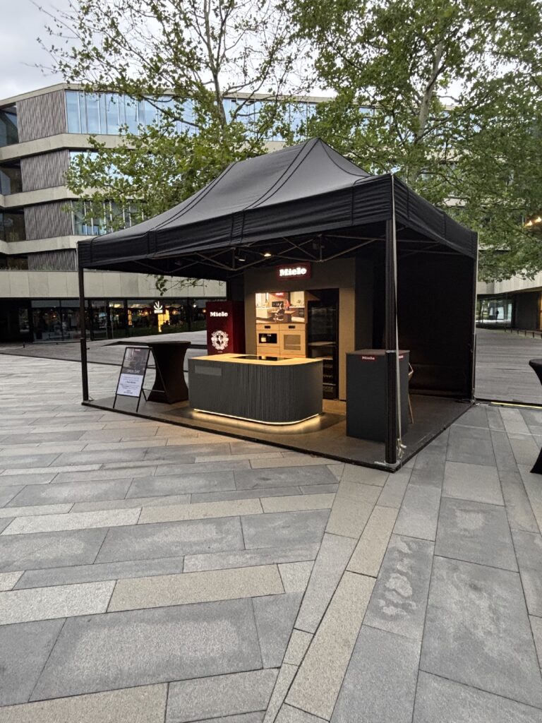 Czarny namiot typu pop-up z logo Miele osłania nowoczesny, zakrzywiony blat, na którym wyeksponowano sprzęt kuchenny, na miejskim placu w pobliżu Royal Wilanów. Ta elegancka konstrukcja idealnie wtapia się w pustą przestrzeń, a w tle widać drzewa, nowoczesne budynki i szarą kamienną nawierzchnię. projekt i nadzór Tatjana Kobuszewska