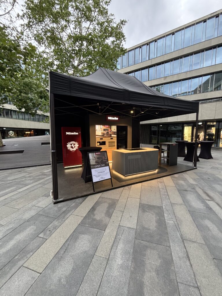 Na miejskim placu z szarą kamienną nawierzchnią w pobliżu Królewskiego Wilanowa rozstawiono nowoczesny, czarny namiot typu pop-up firmy Miele. W środku znajdują się podświetlane ekspozycje i lada, a na zewnątrz – szyld w kształcie litery A. W tle zabudowę uzupełniają budynki biurowe i drzewa. Łącznie: 270 znaków. projekt i nadzór Tatjana Kobuszewska